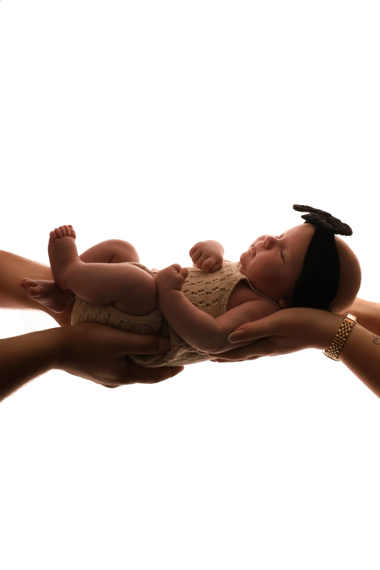 Slapende pasgeboren baby met een zwart strikje, gedragen op de handen van beide ouders in zacht tegenlicht