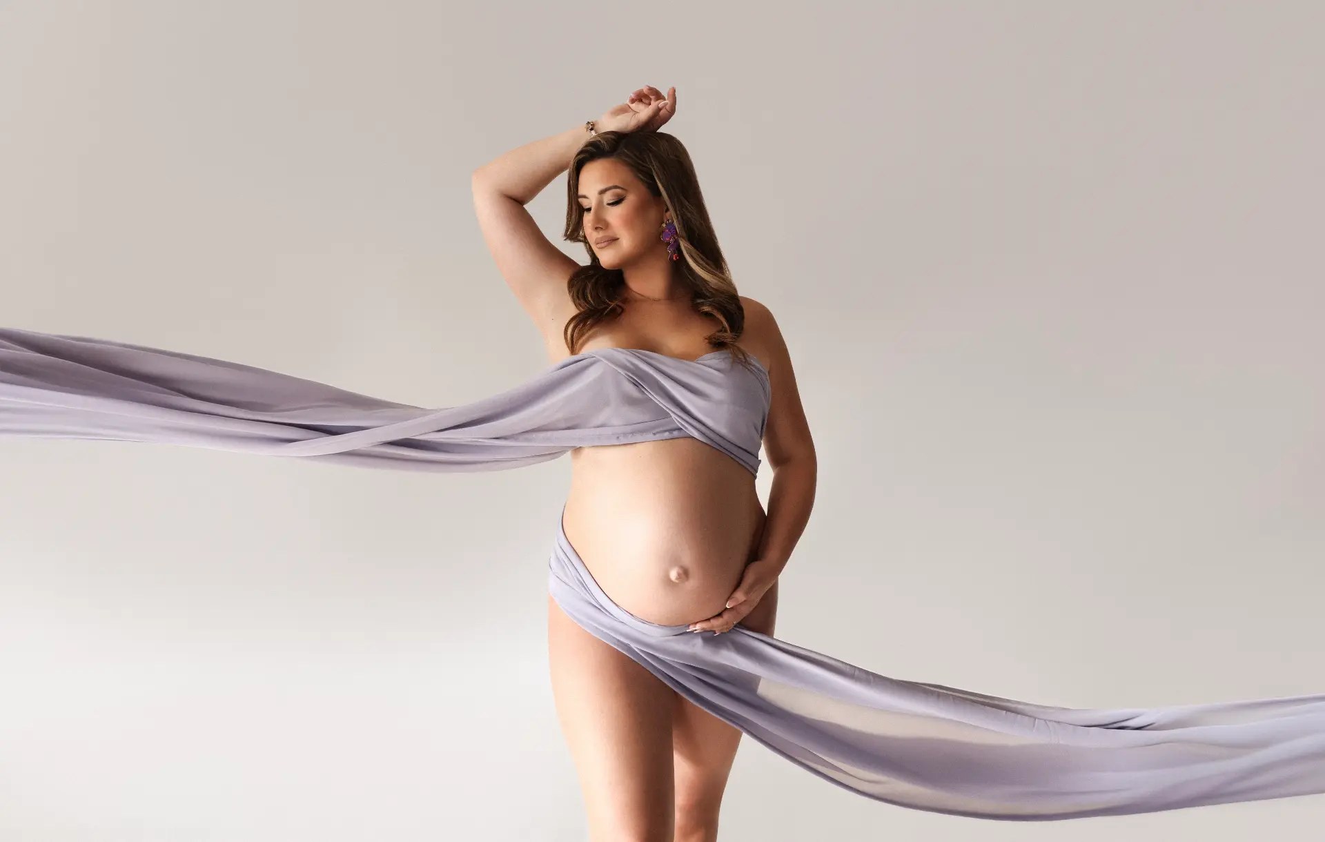 Zwangere vrouw in een lavenderkleurige stof, in de studio, met een hand bij de buik